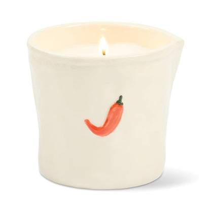A glowing white Piment - Bistro 8 oz Candle sits in a ceramic holder, adorned with a charming small red chili pepper illustration on the front.