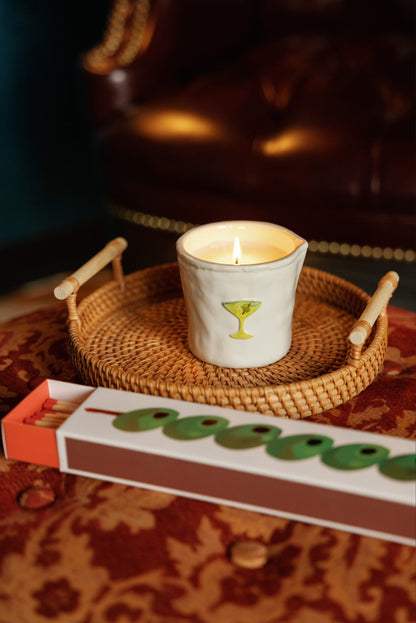 The Martini - Bistro 8 oz Candle, in a white holder featuring a martini glass design, sits on a woven tray beside a box of green-tipped matches atop a patterned surface, with a brown leather chair in the background.