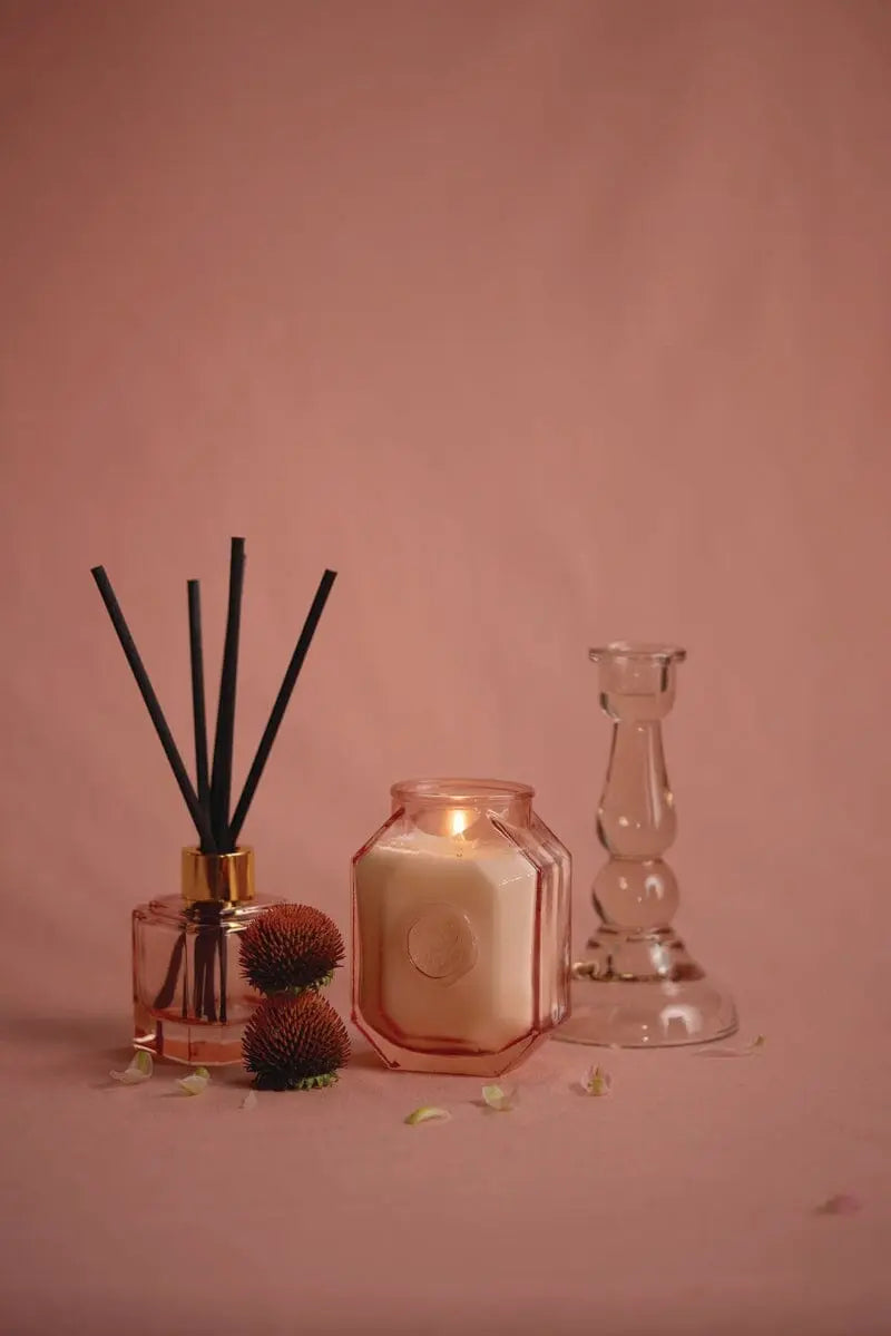 A stylish display featuring the Saffron Rose - Botanica Diffuser 2.88 oz from the luxurious Botanica Collection, paired with a lit candle, dried flowers, and a clear glass holder on a soft pink backdrop.