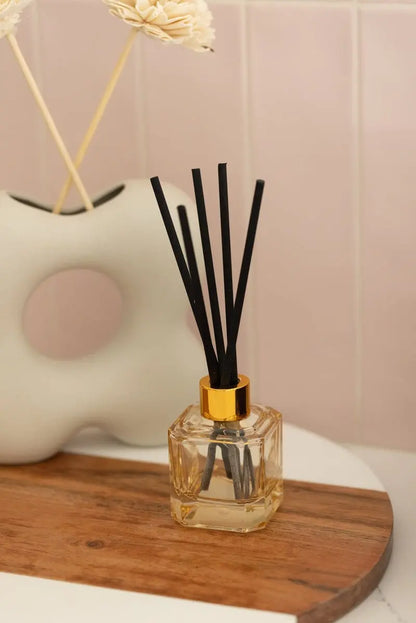 The Cotton + Teak - Botanica Diffuser 2.88 oz, featuring black reeds and a gold cap, is displayed on a wooden tray with a white vase and set against a pink tiled background.