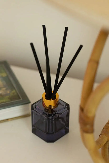 The Sea Salt + Amber - Botanica Diffuser 2.88 oz with black reeds sits on a white surface beside a closed book and rattan chair arm, filling the space with a modern, serene fragrance from the Luxurious Botanica Collection.
