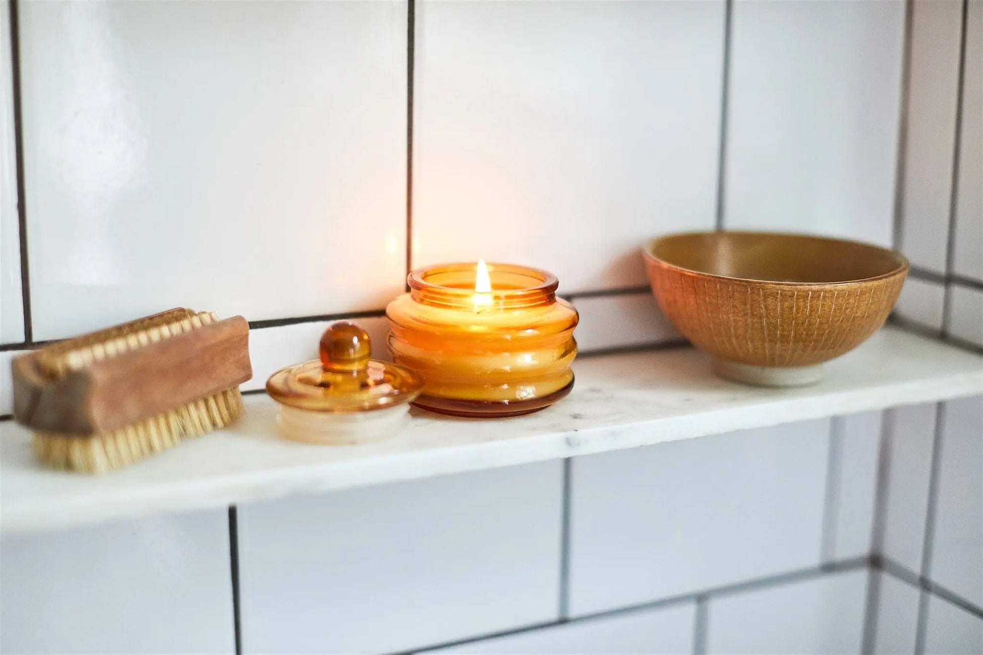 The Meyer Lemon - Beam 3 oz Candle glows beside a small wooden brush, a lidded jar, and a brown bowl, all artfully arranged on a marble shelf against a white tiled wall.