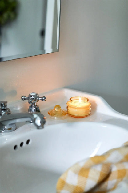 The Meyer Lemon - Beam 3 oz Candle in a yellow glass container adds ambient light to the bathroom, resting on a white sink next to a silver faucet and a yellow checkered cloth.