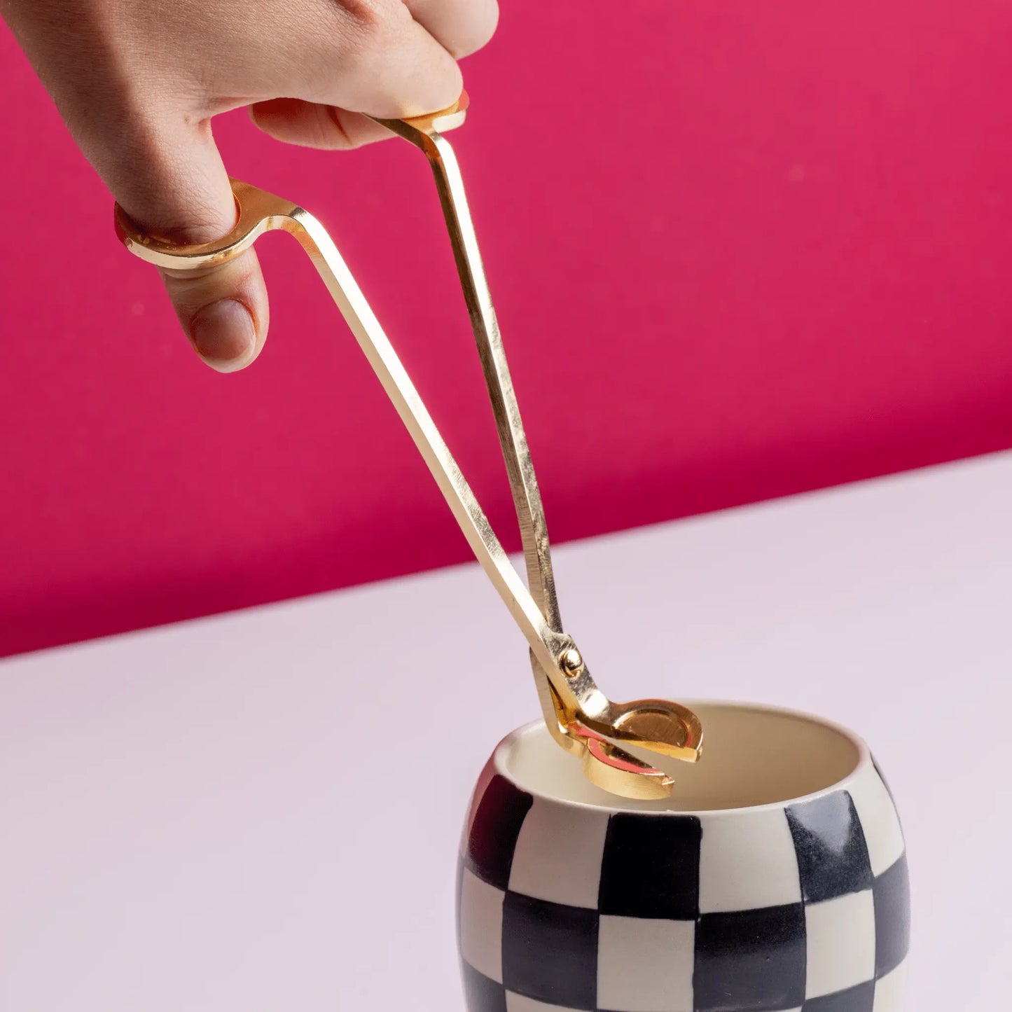 A hand holds the Wick Trimmer above a black-and-white checkered cup, set against a pink background on a white surface—a stylish candle accessory scene.