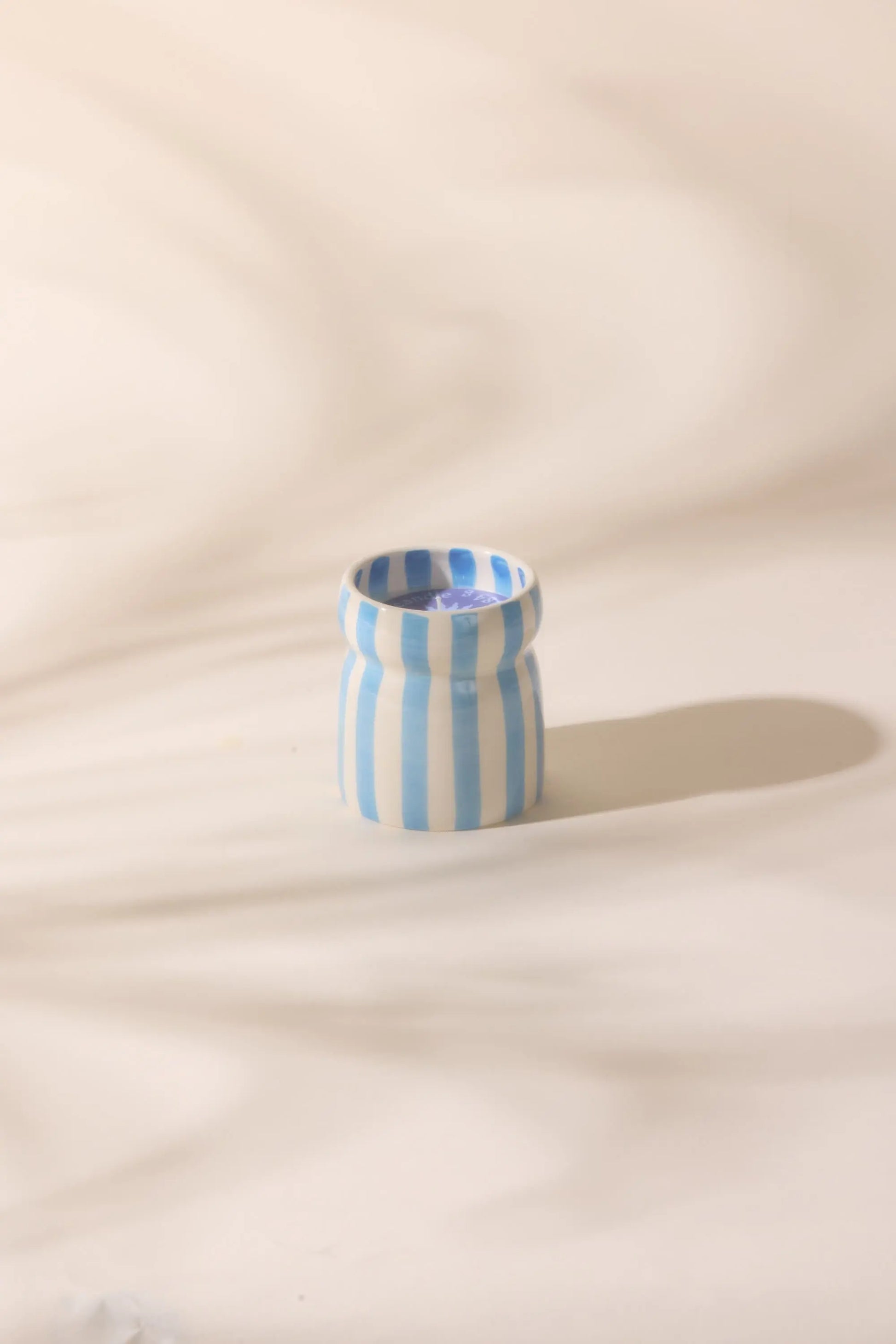 A small ceramic vase with vertical blue and white stripes rests on a light surface, evoking the coastal charm and refreshing ocean tide scent of the Lost At Sea - Cabana 6.5 oz Candle.