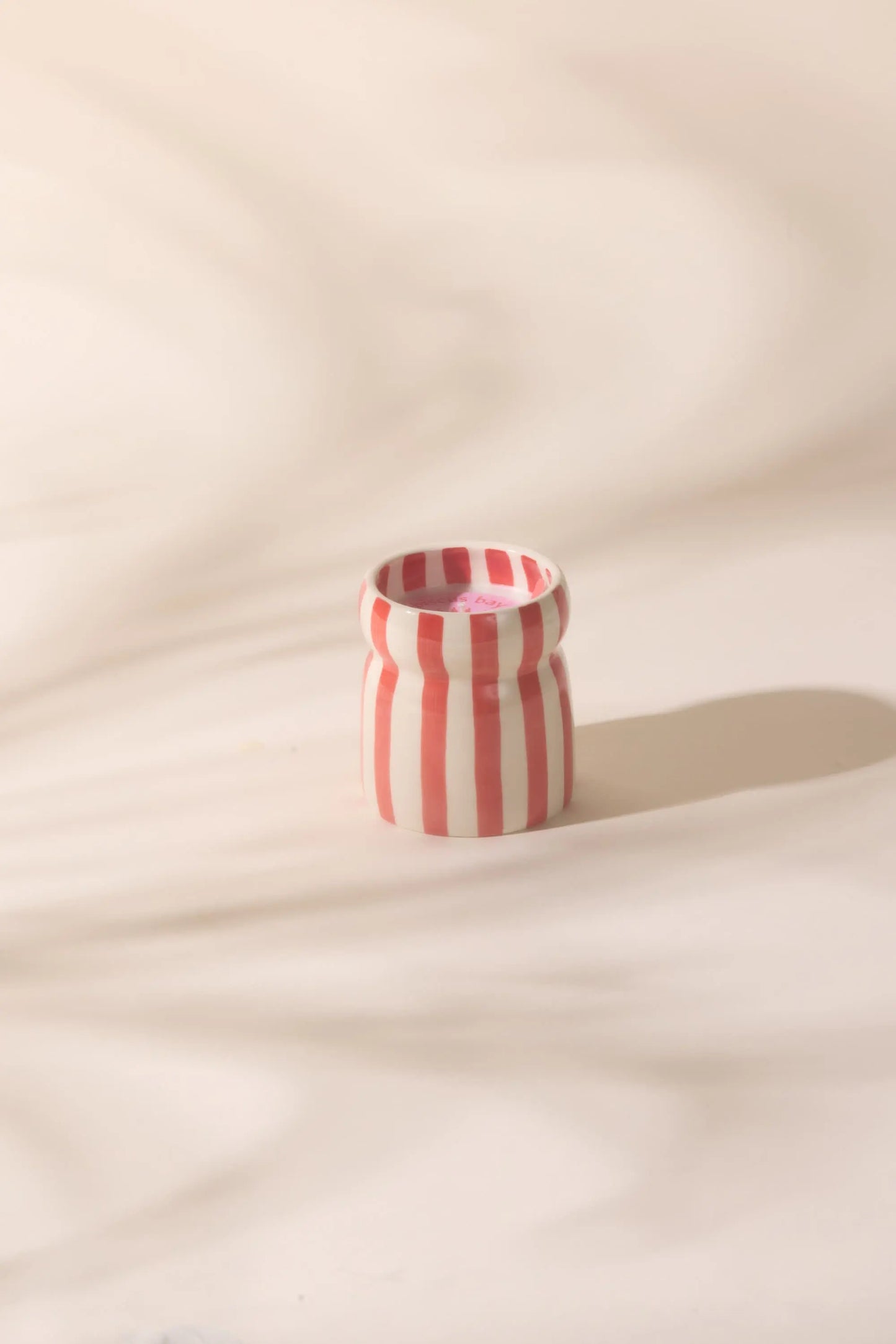 The Hibiscus Bay - Cabana 6.5 oz Candle, a small ceramic vessel with vertical red and white stripes, rests on a light surface, evoking coastal scents in gentle, diffused light.