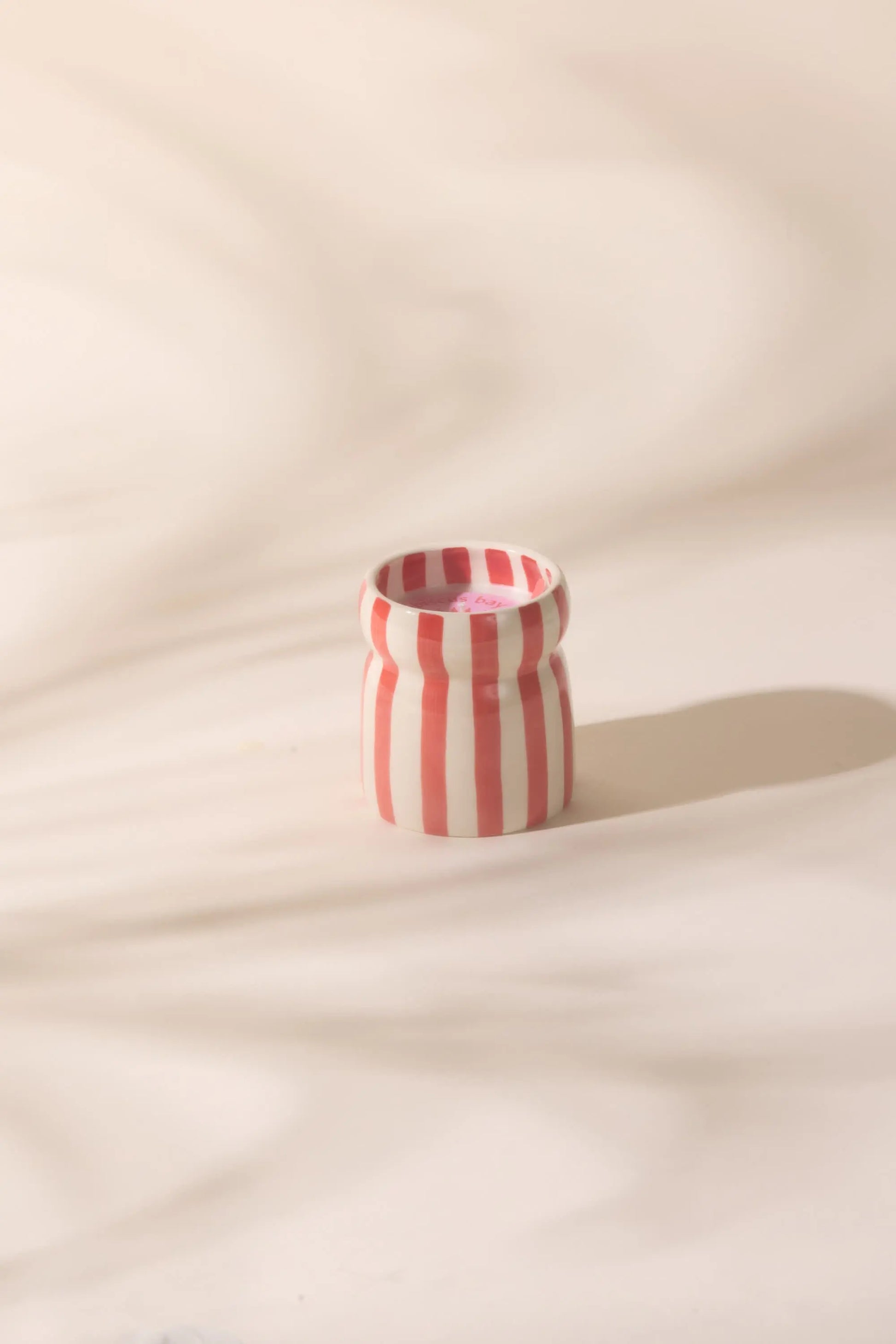 The Hibiscus Bay - Cabana 6.5 oz Candle, a small ceramic vessel with vertical red and white stripes, rests on a light surface, evoking coastal scents in gentle, diffused light.