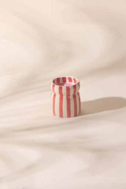 The Hibiscus Bay - Cabana 6.5 oz Candle, a small ceramic vessel with vertical red and white stripes, rests on a light surface, evoking coastal scents in gentle, diffused light.