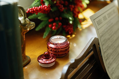 The Winter Berry - Beam 3 oz Candle, with its red and glittery holder, glows beside sheet music and festive greenery with red berries on a wooden surface—creating a cozy holiday vibe that's perfect for gifting.