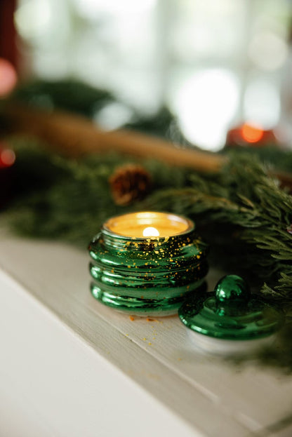 The Balsam & Fir - Beam 3 oz Candle in a green glass jar sits on a white surface with its lid, surrounded by pine branches and holiday decor—creating a cozy, festive vibe that's perfect as a stocking stuffer or Christmas party favor.