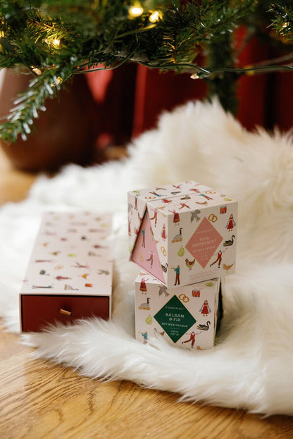 The Pink Peppermint - 12 Days Of Christmas gift boxes, featuring festive holiday artwork, sit on a white fur rug under a lit Christmas tree. One box is open, showcasing cheerful seasonal designs perfect for celebrating the holidays.