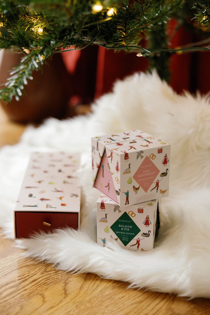 Two decorative gift boxes sit on a white faux fur rug under a Christmas tree, beside a rectangular Balsam & Fir - 12 Days Of Christmas box. Warm lighting and greenery create a cozy holiday atmosphere.