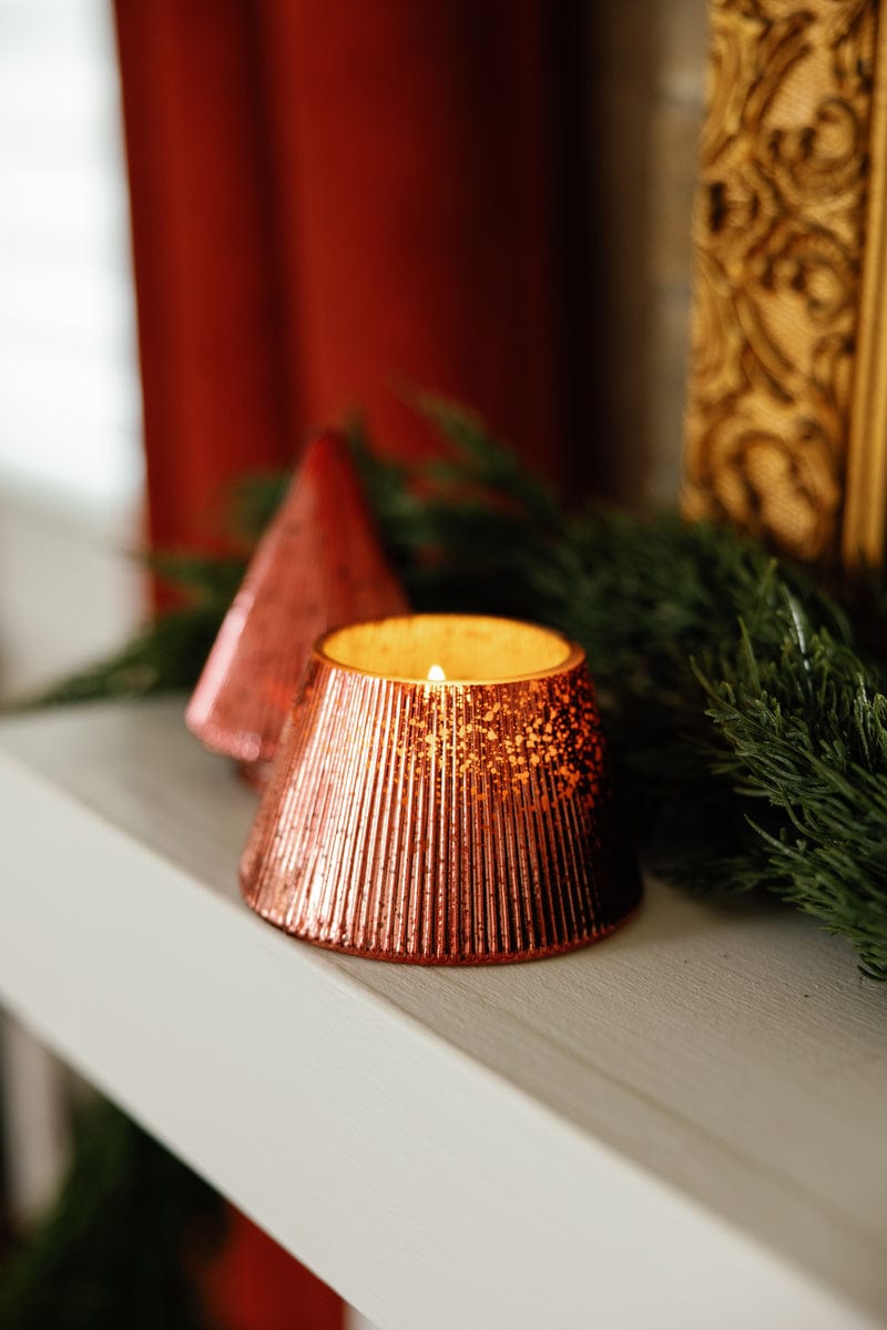 A lit Winter Berry Tree - 7 oz Candle in a ribbed metallic holder with a copper finish sits on a white shelf, accented by evergreen garland and another matching mercury glass candle in the background.