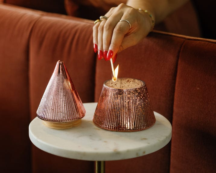 A person with red nails lights a Pink Peppermint Tree - 7 oz Candle in a mercury glass holder on a round white table by a brown sofa.