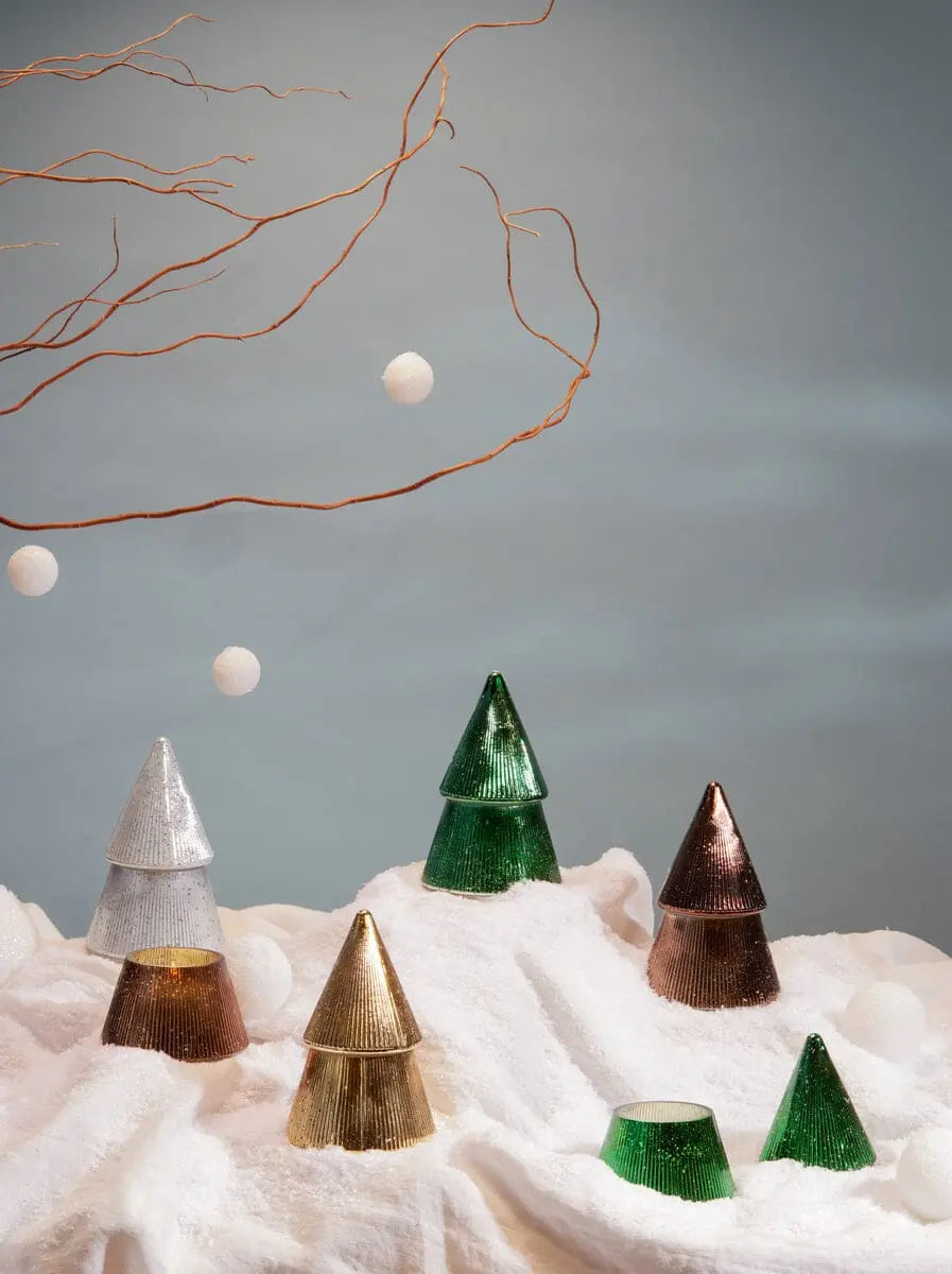 Green, silver, and gold tree-shaped glass ornaments rest on a snowy surface. Above them, bare branches and floating white balls are suspended against a soft blue-gray background. The shimmering mercury glass finish of the ornaments recalls the nostalgic glow of the Cypress + Fir Green Tree - 7 oz Candle.