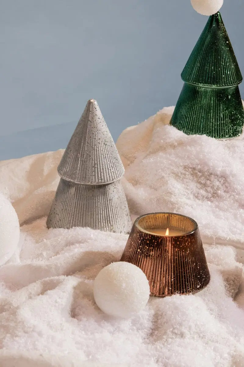 The Cypress + Fir Silver Tree - 7 oz Candle sits on artificial snow with white snowballs, creating a festive winter holiday scene against a soft blue background.