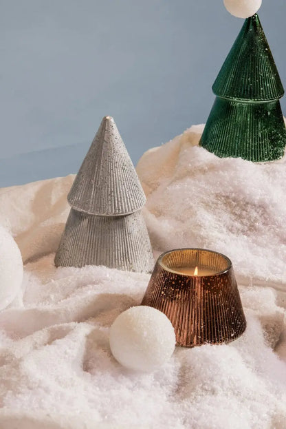 The Cypress + Fir Silver Tree - 7 oz Candle sits on artificial snow with white snowballs, creating a festive winter holiday scene against a soft blue background.