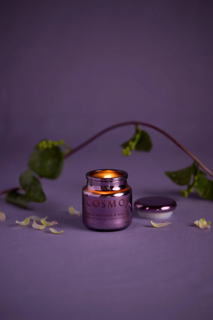 A Wisteria & Willow - Chroma Candle 2.5oz in an electroplated glass jar rests on a purple surface with flower petals and a vine nearby; the lid lies beside it, reflecting Chroma candles' Latin-inspired fragrance.
