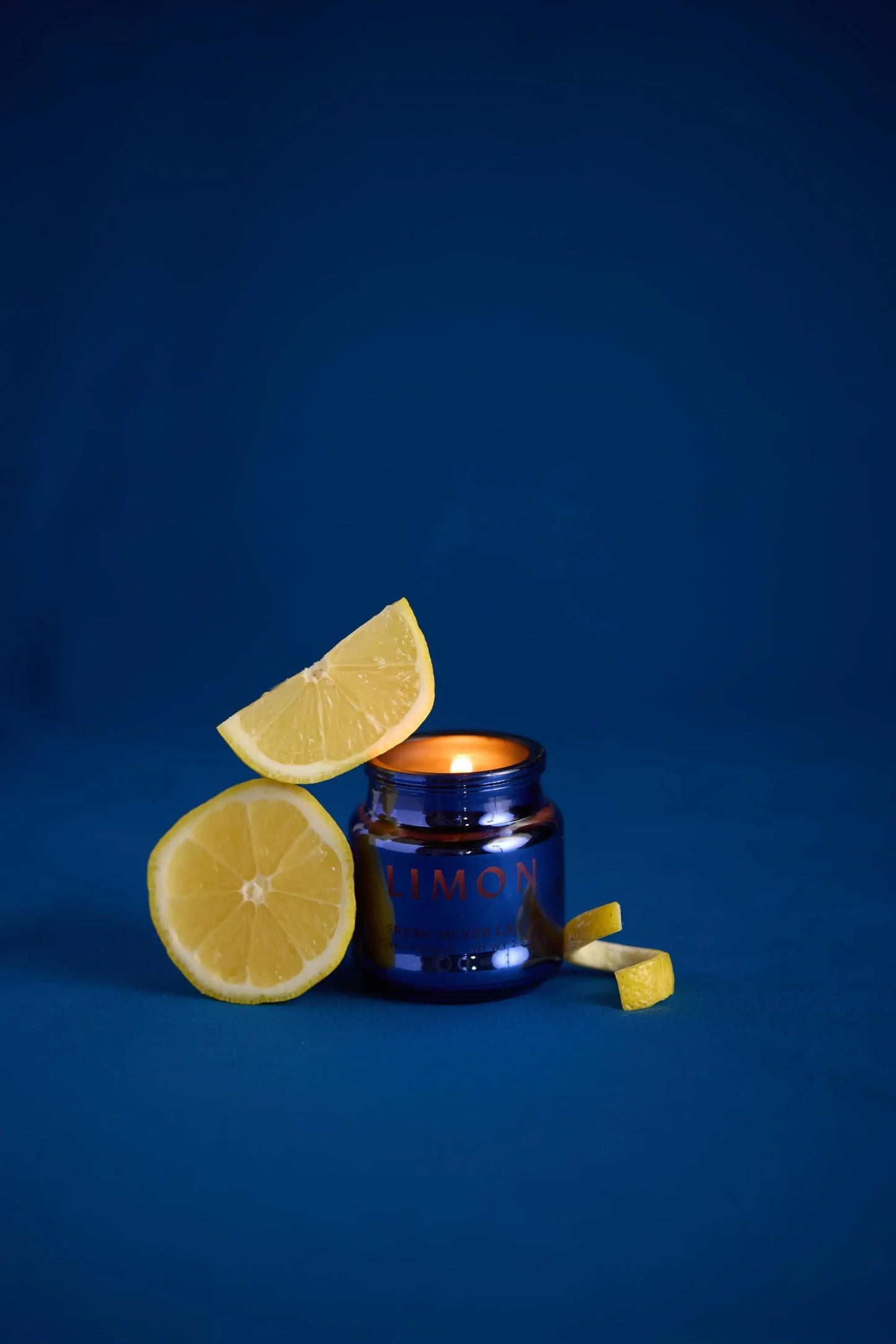 A lit Meyer Lemon - Chroma Candle 2.5 oz in a blue glass jar sits among lemon wedges and a half lemon, set against a solid blue background.