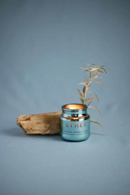 A lit Sea Salt & Sage - Chroma Candle 2.5 oz sits beside driftwood and a dried plant against a plain blue background.