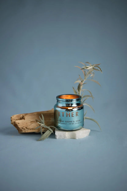 A blue Sea Salt & Sage - Chroma Candle 2.5 oz rests on a white stone, with a piece of wood and silvery foliage beside it, set against a blue background.