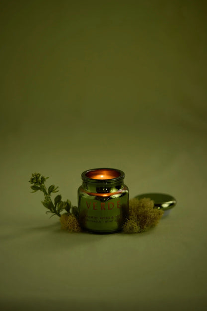 A lit Pacific Moss & Mist - Chroma Candle 2.5 oz jar sits on a light surface, surrounded by moss and a plant sprig, with its metallic lid beside it.