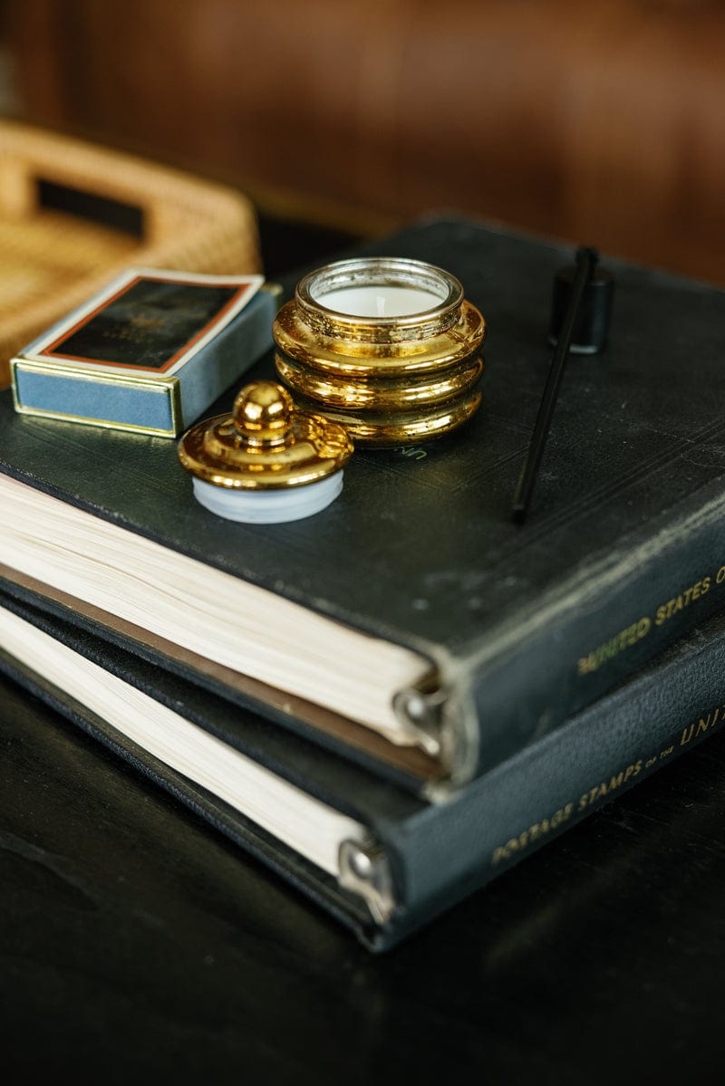 A close-up of two stacked black books with gold titles, a vintage gold ink pot, black dip pen, matches, and the Cypress & Fir - Beam 3 oz Candle—perfect as a gift-ready stocking stuffer or paired with your favorite holiday candle.