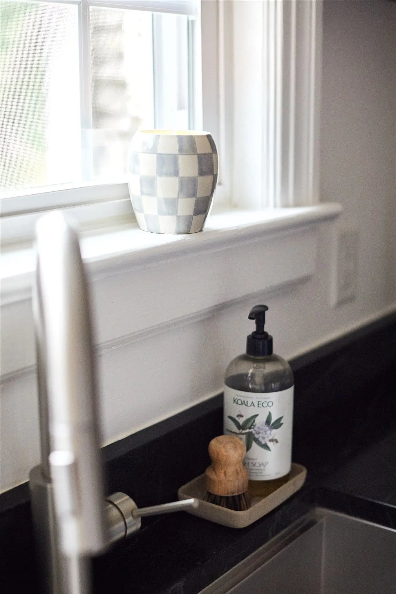 A checkered cup sits on a windowsill above the kitchen sink. On the counter, a Koala Eco hand wash and scrub brush in a tray complement reusable decor like the Cotton + Teak Checkmate Candle beside the faucet.