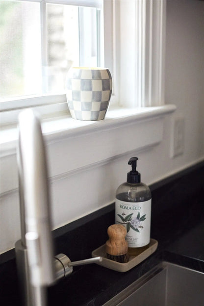 A checkered cup sits on a windowsill above the kitchen sink. On the counter, a Koala Eco hand wash and scrub brush in a tray complement reusable decor like the Cotton + Teak Checkmate Candle beside the faucet.