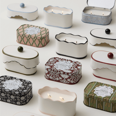 Assorted decorative candles in patterned, rectangular tins arranged on a white surface; some tins are open, showing lit candles inside.