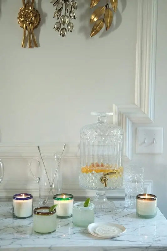 An Amber + Coconut - La Playa 9 oz Candle sits on a marble countertop among artisan Mexican candles, hand-blown bubble glassware, a glass drink dispenser with citrus slices, and gold wall decor.