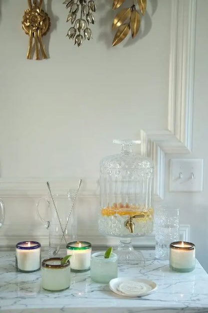 An Amber + Coconut - La Playa 9 oz Candle sits on a marble countertop among artisan Mexican candles, hand-blown bubble glassware, a glass drink dispenser with citrus slices, and gold wall decor.