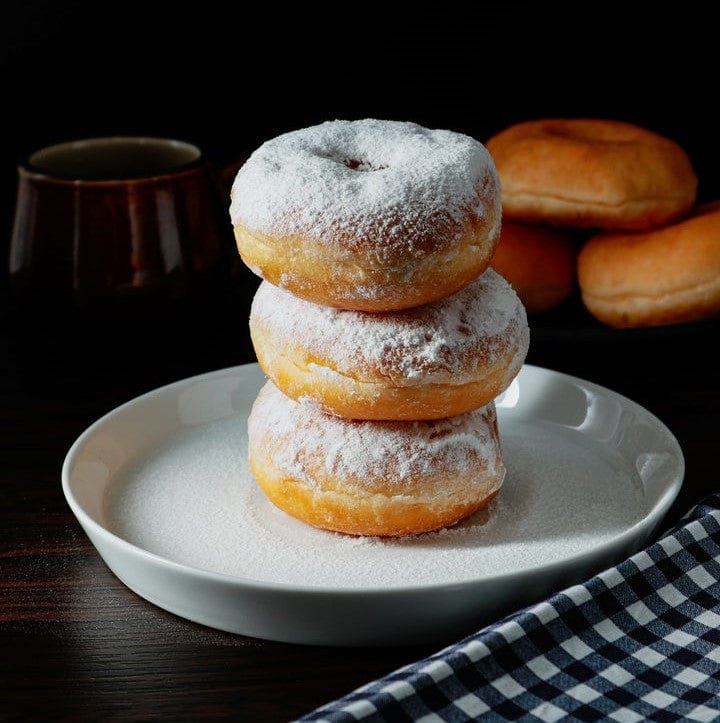 Three powdered donuts are stacked on a white plate, dusted with powdered sugar. More donuts and a dark cup are in the background, with a blue and white checkered cloth beside the plate on a dark surface.