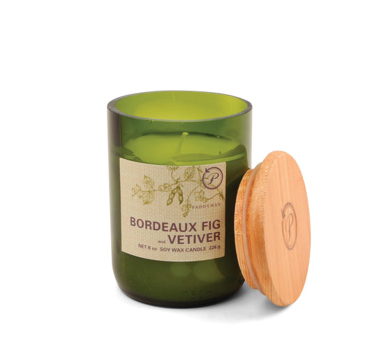 A green recycled glass Bordeaux Fig + Vetiver - Eco 8 oz soy wax candle with a wooden lid rests nearby; its label highlights the scent and eco-friendly features.