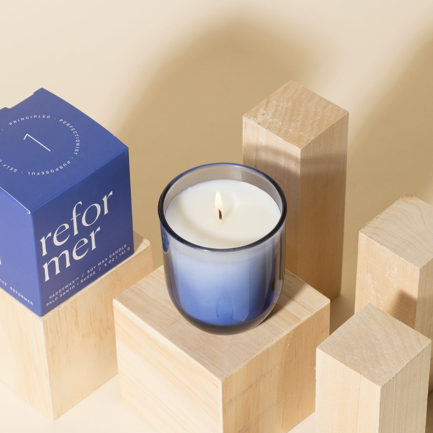A lit white candle in a blue glass holder sits on a wooden block, surrounded by other wooden blocks. A blue box labeled reformer is positioned to the left of the candle. The background is beige.