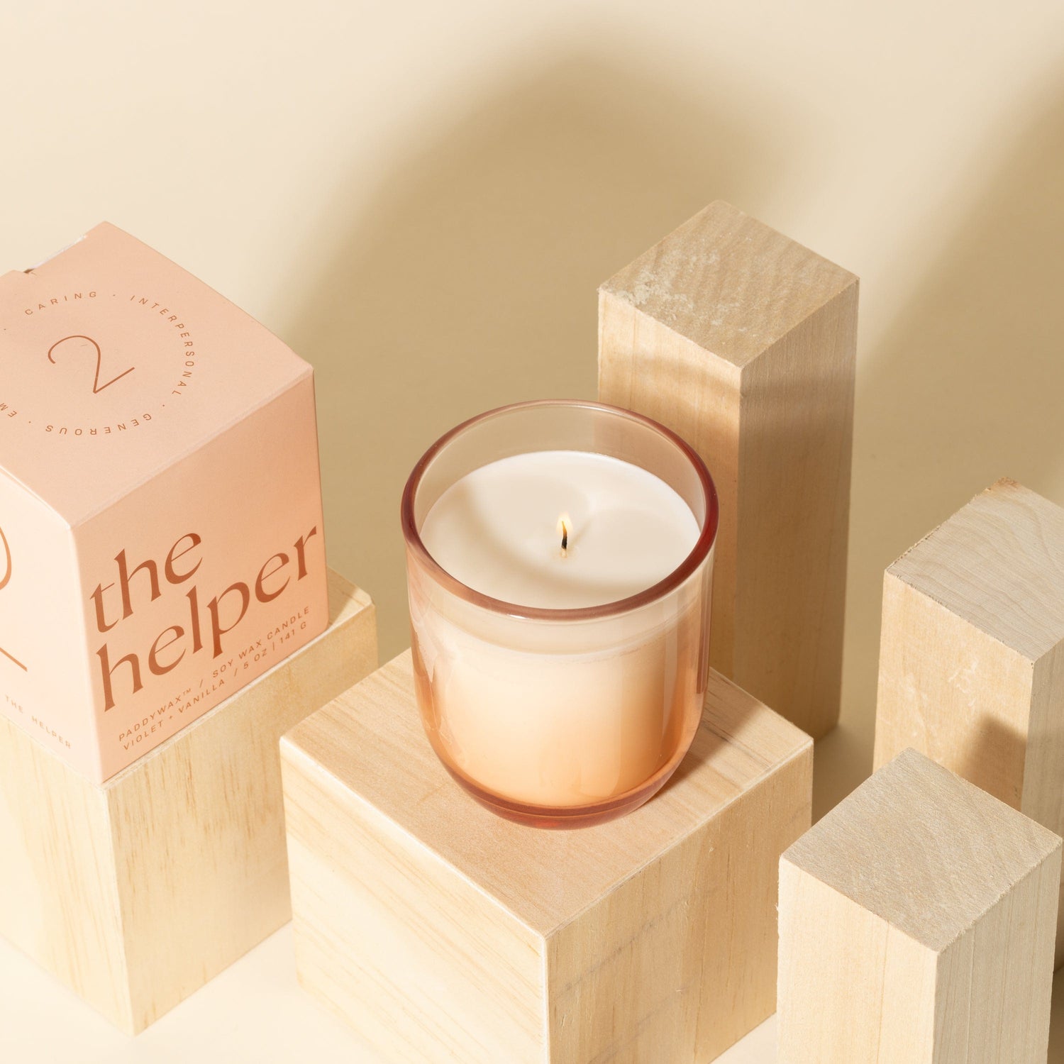 A lit candle in a glass holder sits on a wooden block, surrounded by other wooden blocks. A pink box labeled the helper is positioned next to the candle. The background is a soft beige color.