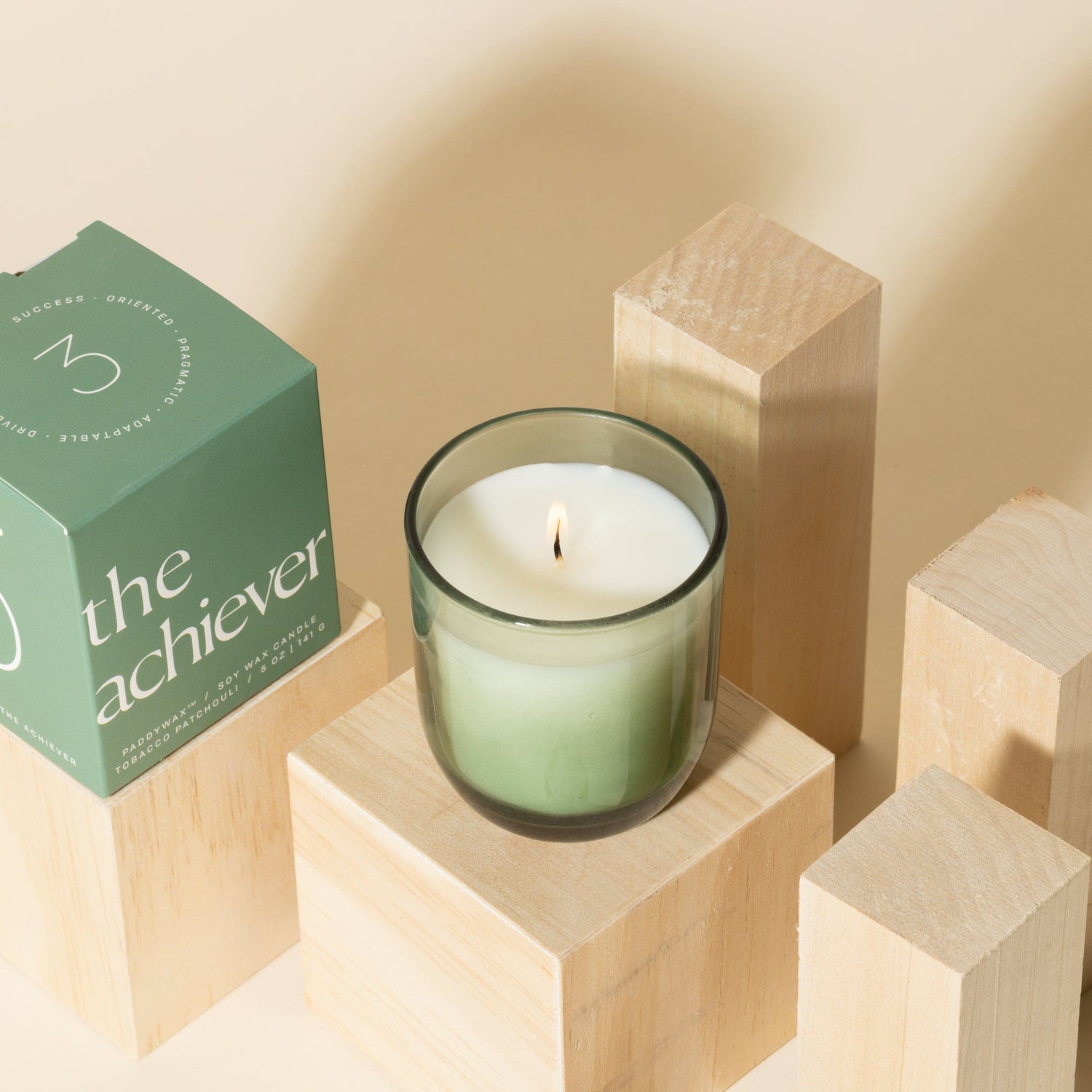 A green glass candle labeled the achiever sits lit on a wooden block beside its matching green box, surrounded by other wooden blocks on a beige background.