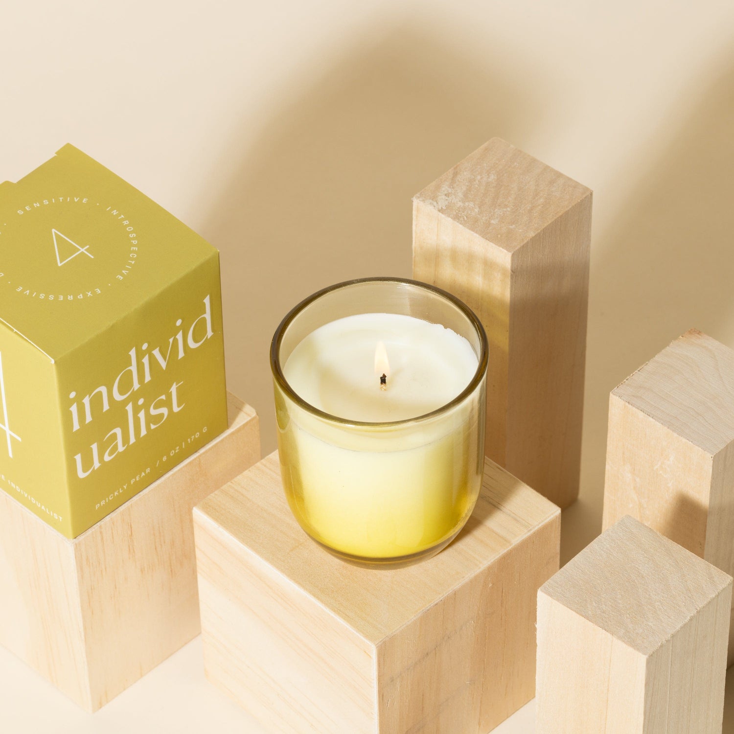 A lit candle in a yellow glass container sits on a wooden block, surrounded by other wooden blocks and a yellow box labeled individualist on a beige background.