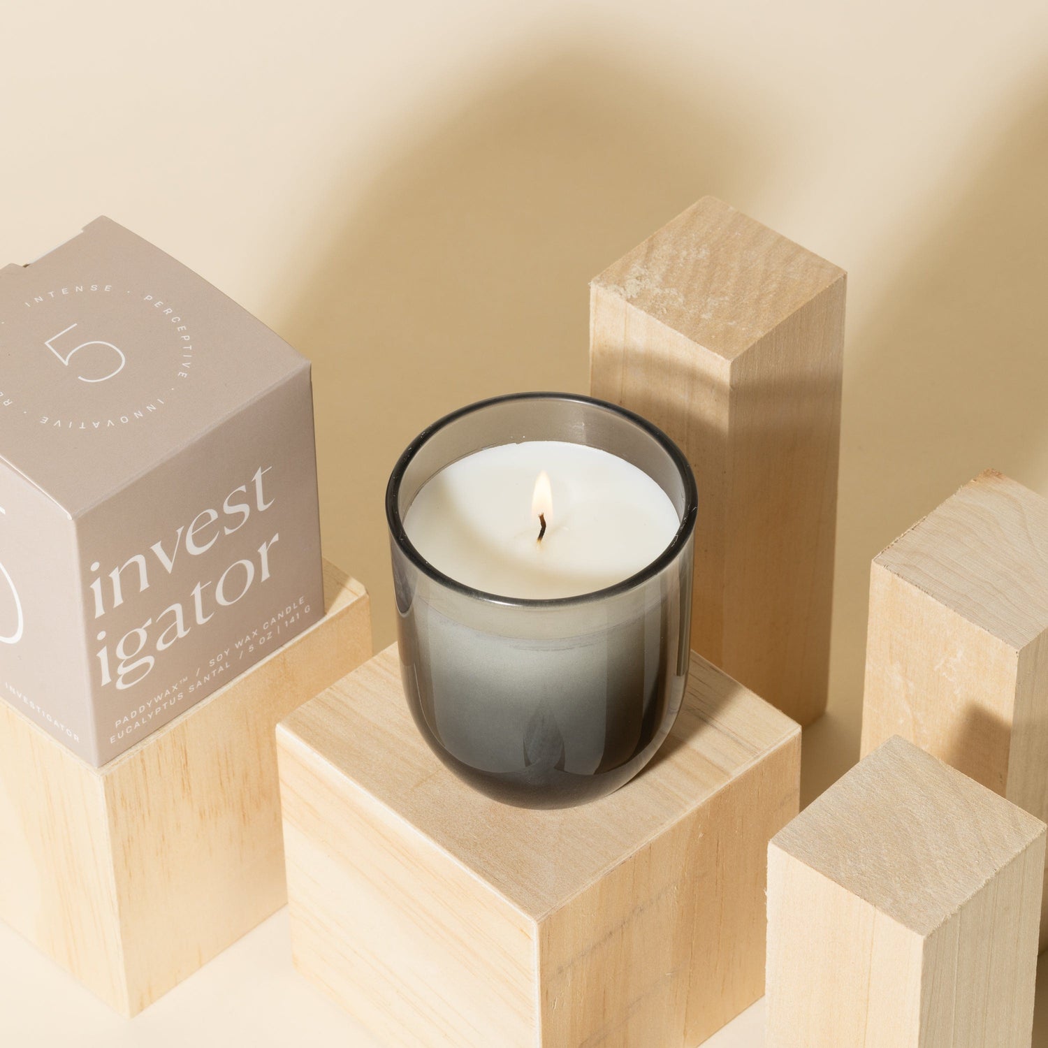 A lit white candle in a gray glass holder sits on wooden blocks next to a beige box labeled investigator on a neutral background.