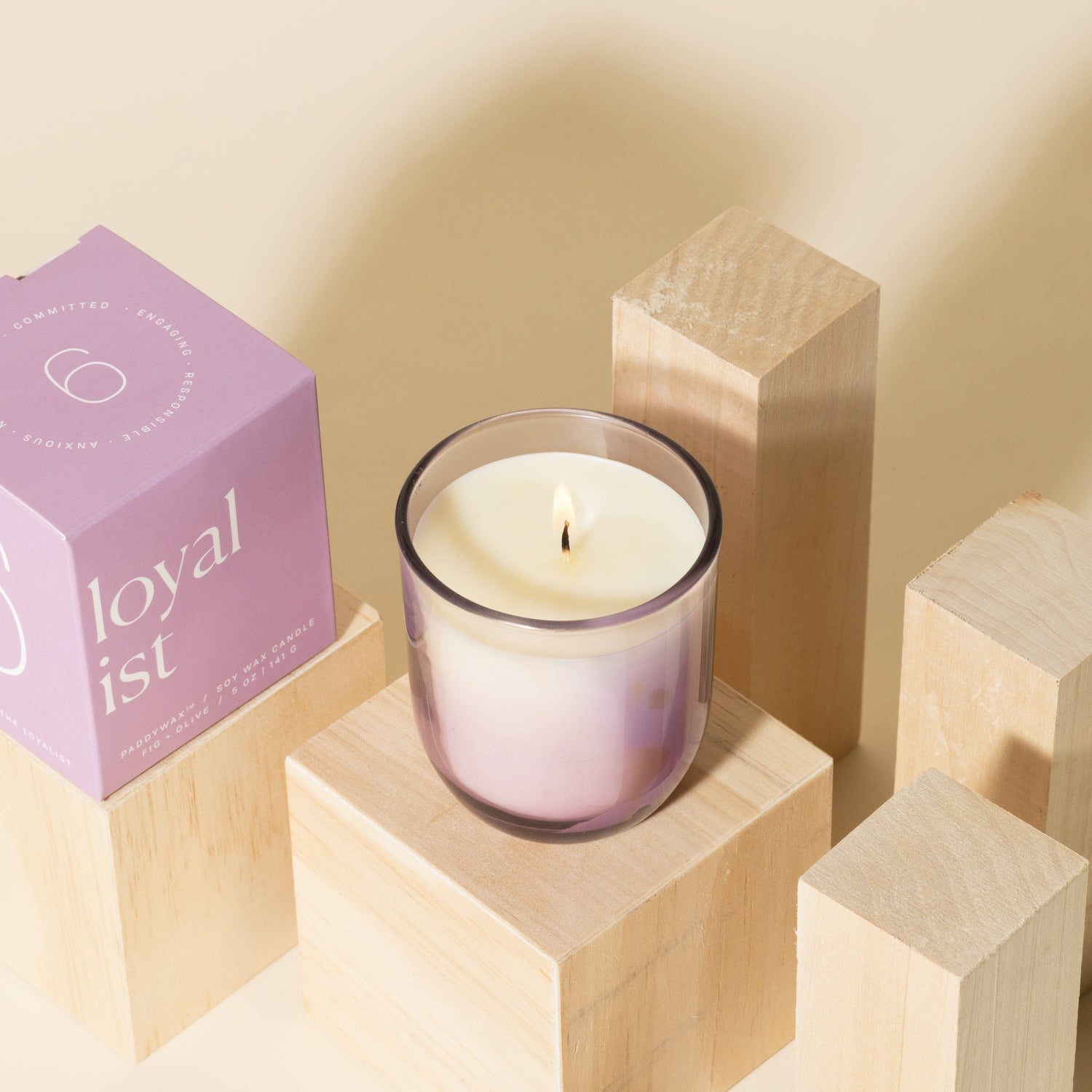 A lit lavender-colored candle in a glass holder sits on wooden blocks next to a purple box labeled loyalist against a beige background.