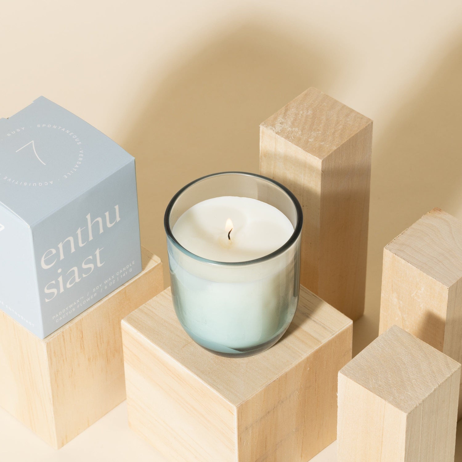 A lit candle in a glass jar sits on a wooden block, surrounded by other wooden blocks. A blue box labeled enthusiast is placed nearby. The background is a soft beige color.