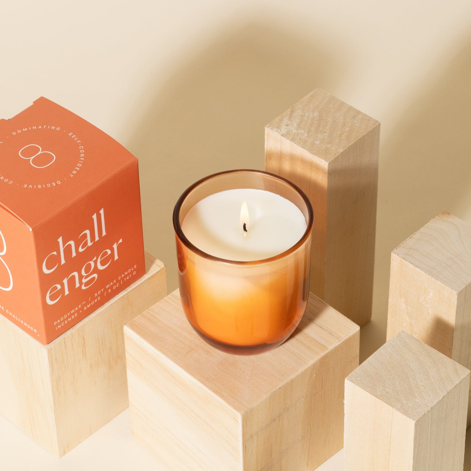An orange glass candle sits lit on a wooden block, next to its orange box labeled challenger, surrounded by additional light wooden blocks against a beige background.