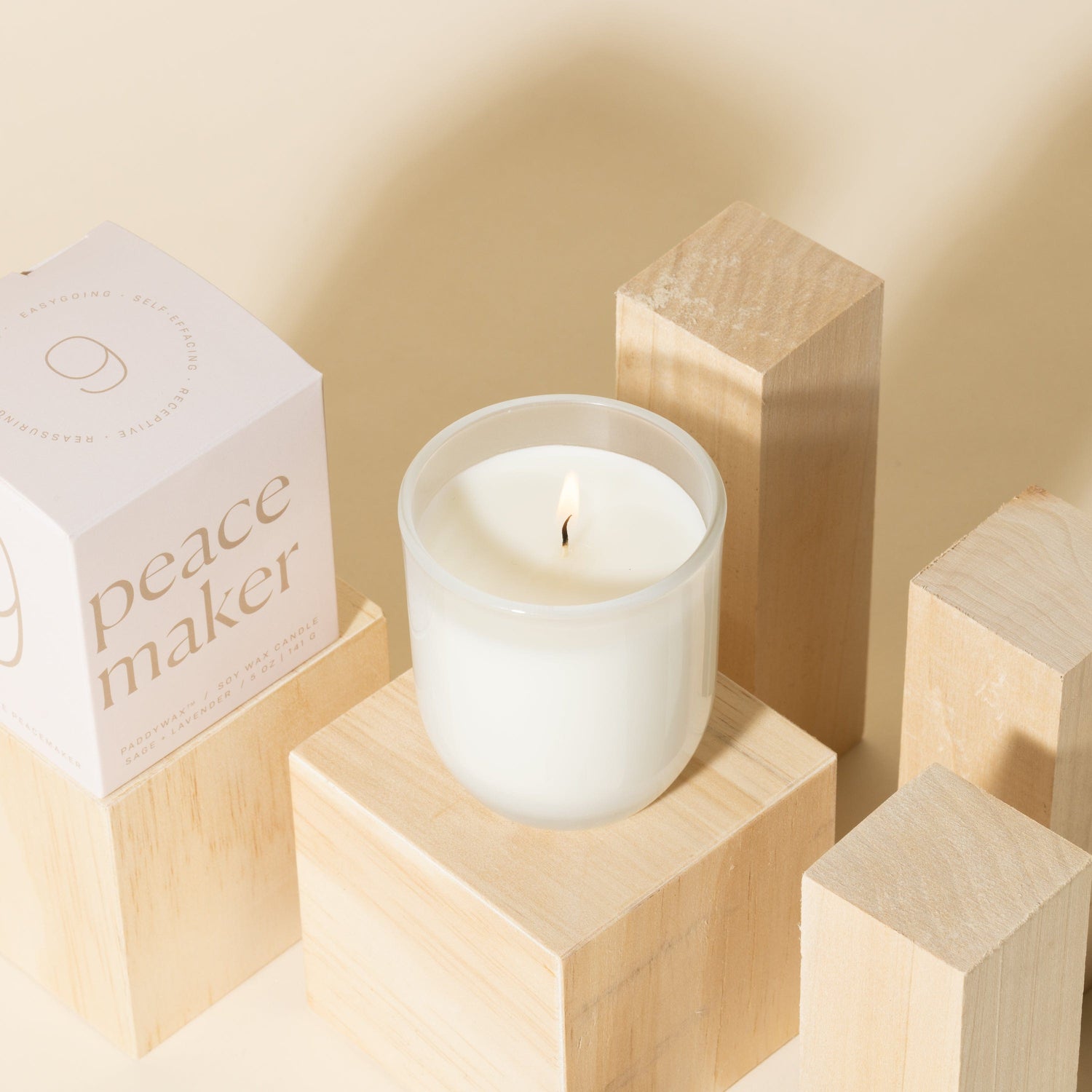 A lit white candle in a glass holder sits on a light wooden block, surrounded by other wooden blocks. A pink box labeled peace maker stands nearby. The background is a soft beige color.