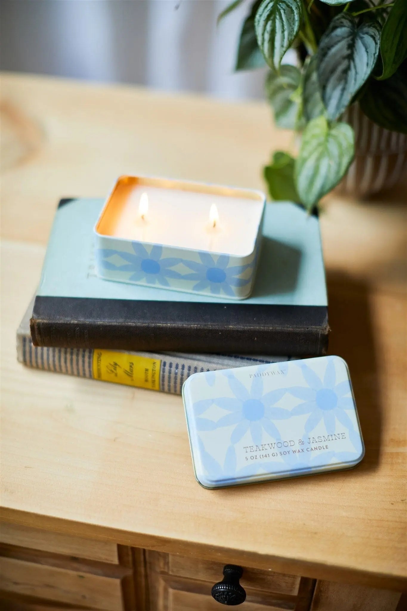 A Teakwood & Jasmine 5 oz Tin Candle with a blue patterned tin sits lit on stacked books next to its lid on a wooden table, with a leafy potted plant nearby.