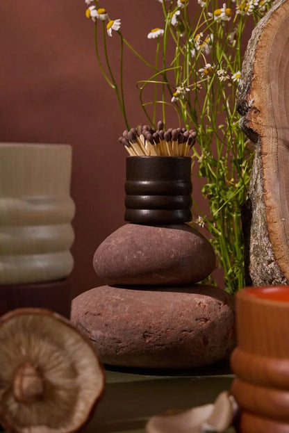 A stack of smooth stones topped with the Dark Brown Ceramic Wood Match Holder, surrounded by flowers, a tree slice with visible woodgrain, and textured ceramic vessels.