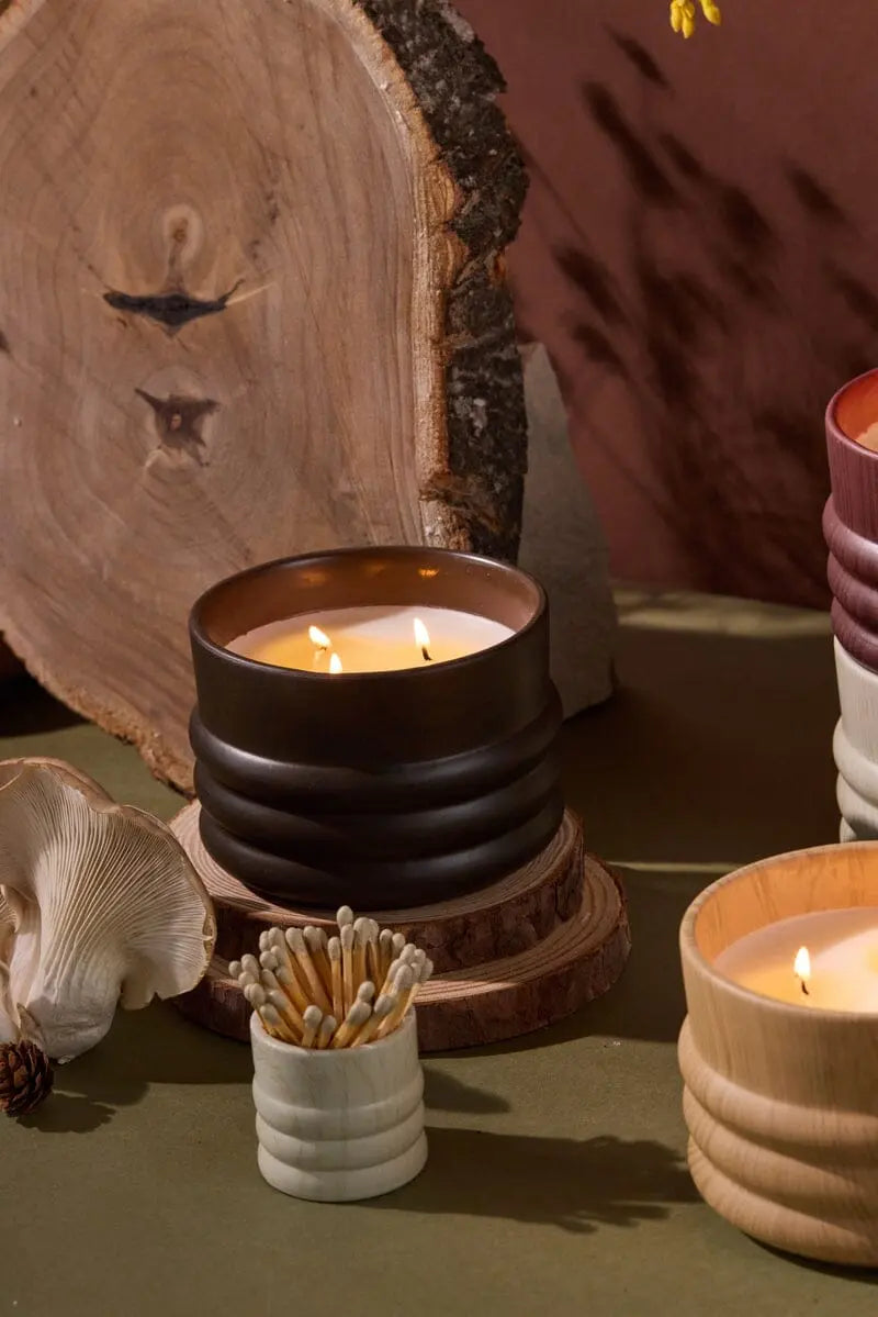 Three lit Tobacco & Patchouli - Grain 12 oz candles in ribbed ceramic holders are displayed on a table with matches, a wooden slab, a mushroom, and a pinecone in the background.