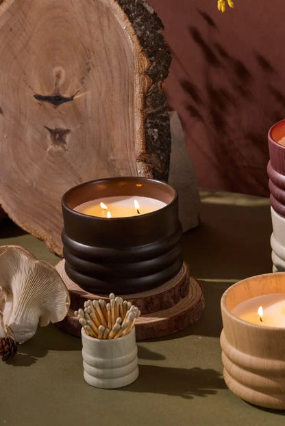 Three lit woodgrain candles in ribbed ceramic holders sit on a green surface near a White Washed Ceramic Wood Match Holder, a wooden log slice, and a mushroom.