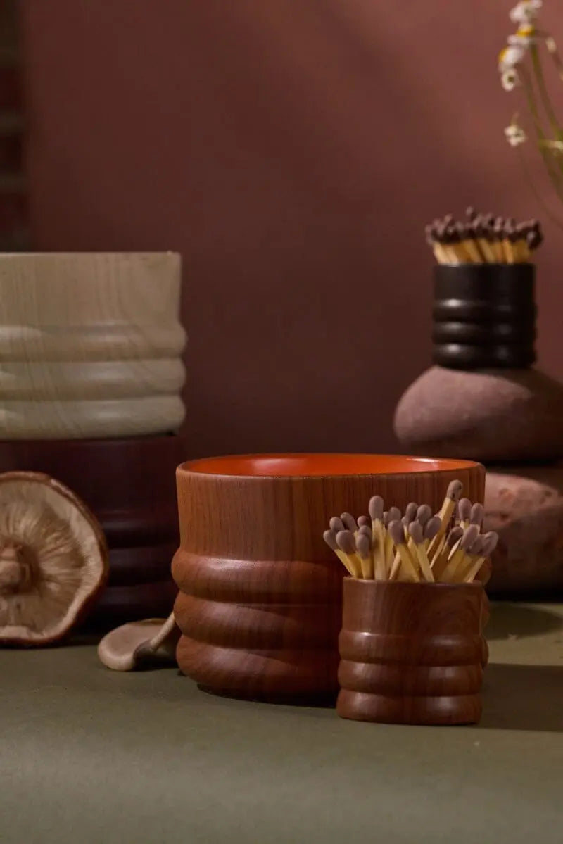 The Redwood & Amber - Grain 12 oz Candle, with elegant woodgrain detail, sits on a green surface among wooden cups and vases, some holding matches, with a dried mushroom and flowers enhancing the warm, natural ambiance.