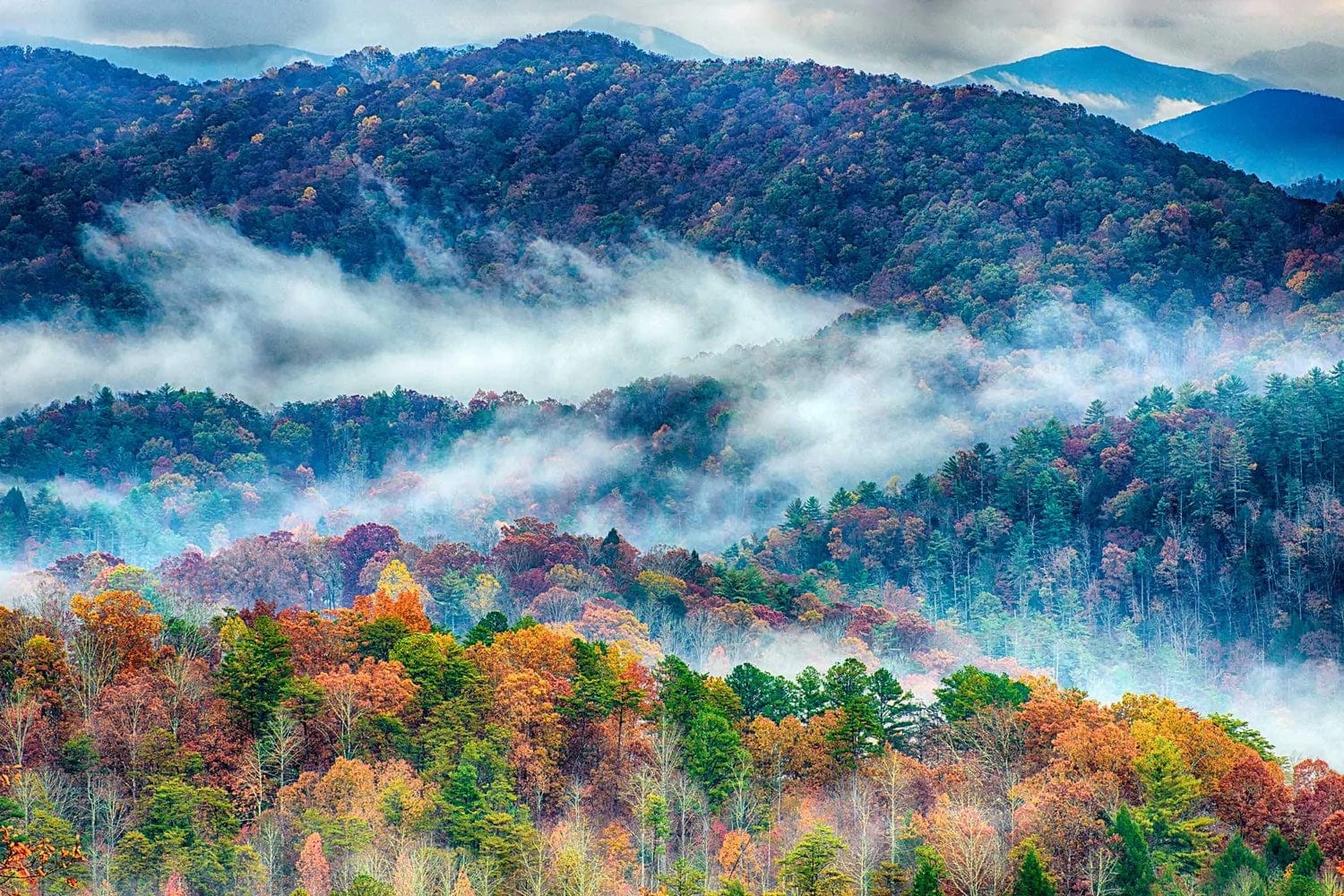 Colorful autumn trees cover rolling hills and mountains, with wisps of fog drifting through the forested landscape under a cloudy sky.