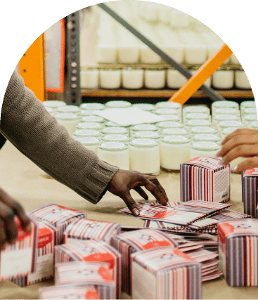 Two people organize striped product boxes on a table, with shelves of glass jars containing white contents in the background.